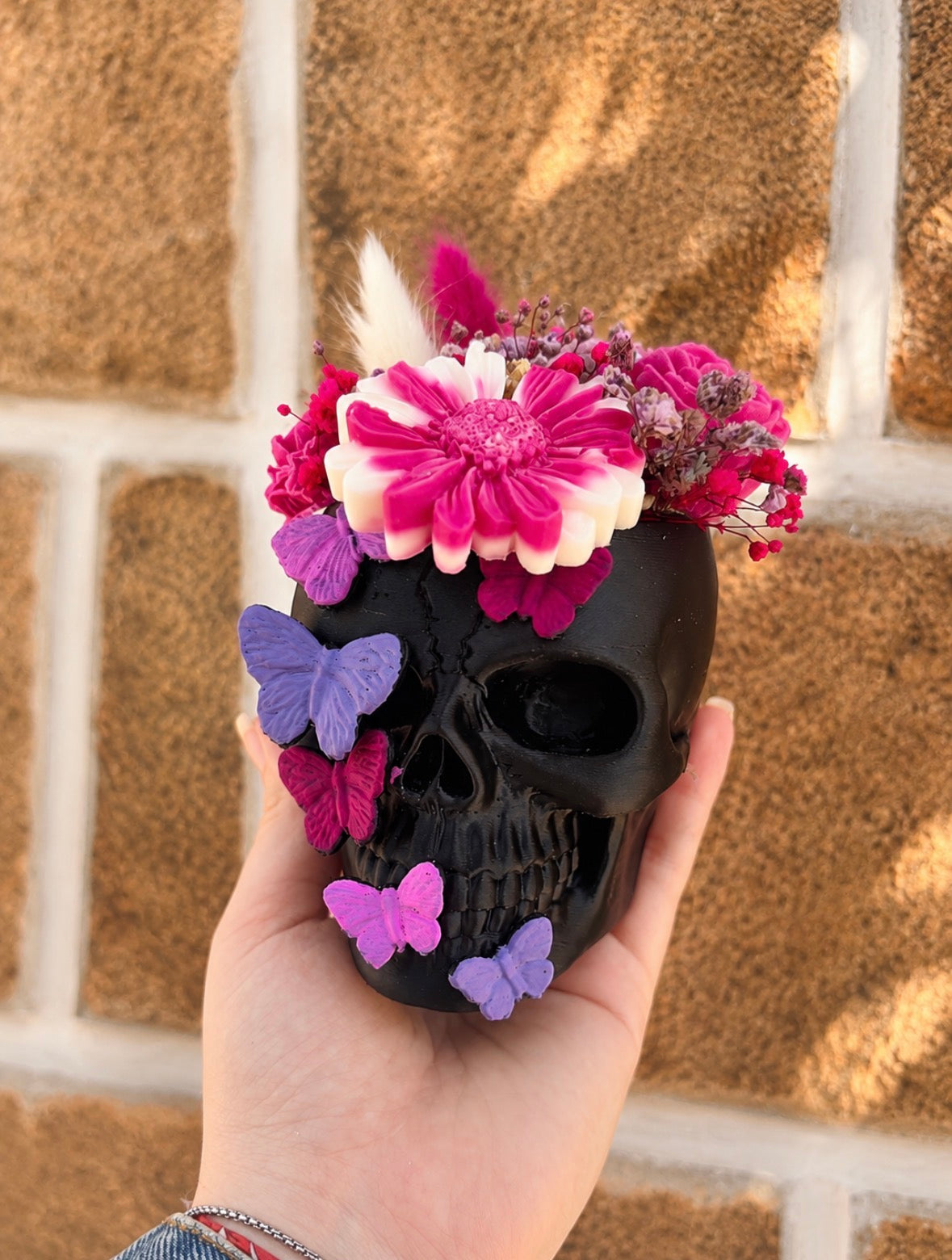 Bouquet dans un crâne noir, orné de fleurs roses et crème et de papillons roses et violets, tenu à la main.